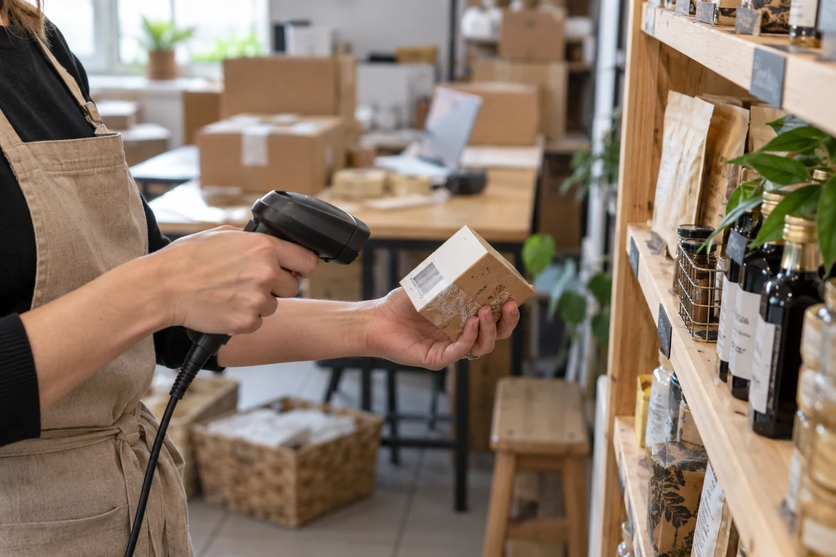 Ruce prodavačky skenující čárový kód malé krabičky u regálu malého obchodu, v pozadí balíky pro e-shop